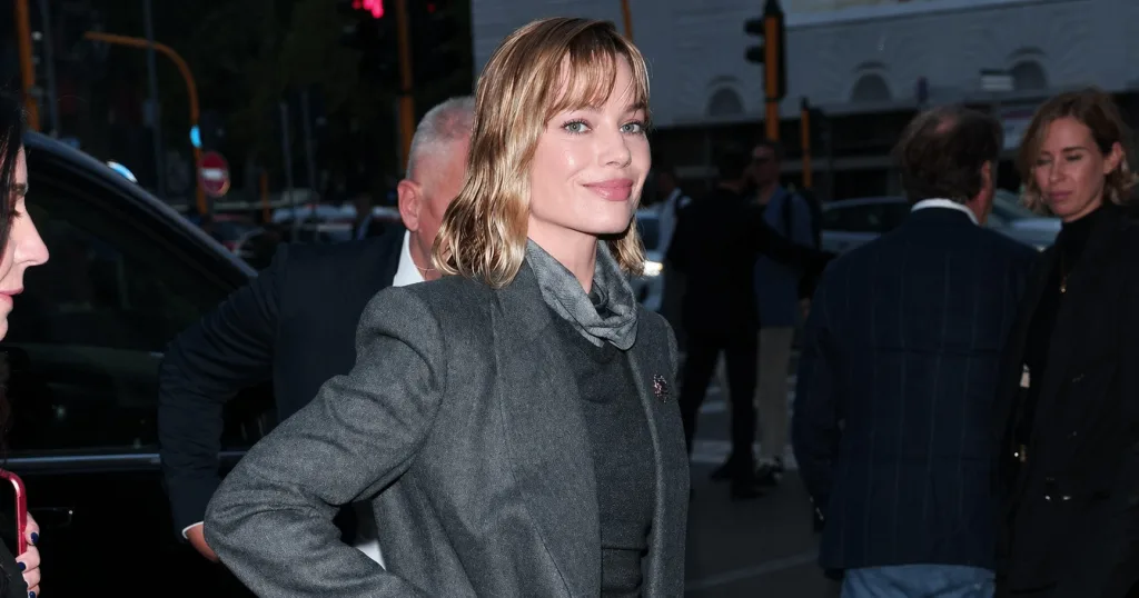 Woman in a gray blazer with a scarf, smiling on a busy city street at dusk.