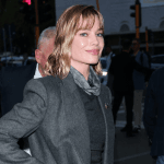 Woman in a gray blazer with a scarf, smiling on a busy city street at dusk.