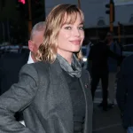 Woman in a gray blazer with a scarf, smiling on a busy city street at dusk.