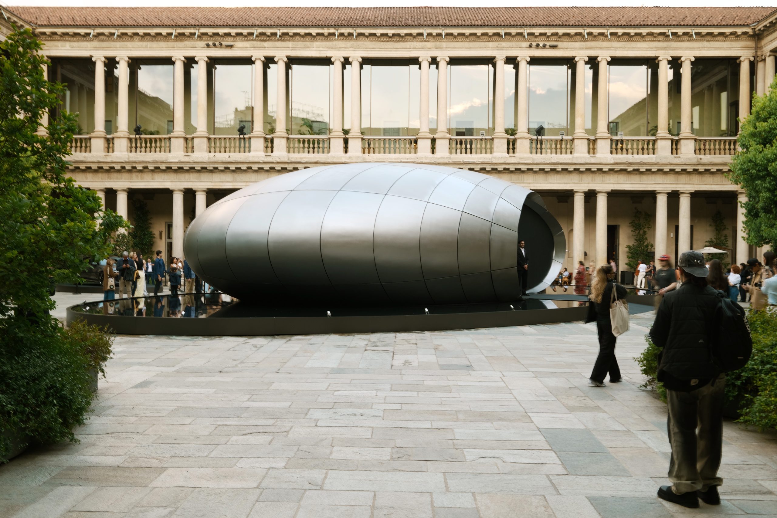 Large silver capsule sculpture in a circular pool, set in a courtyard with a colonnaded building in the background and visitors nearby.
