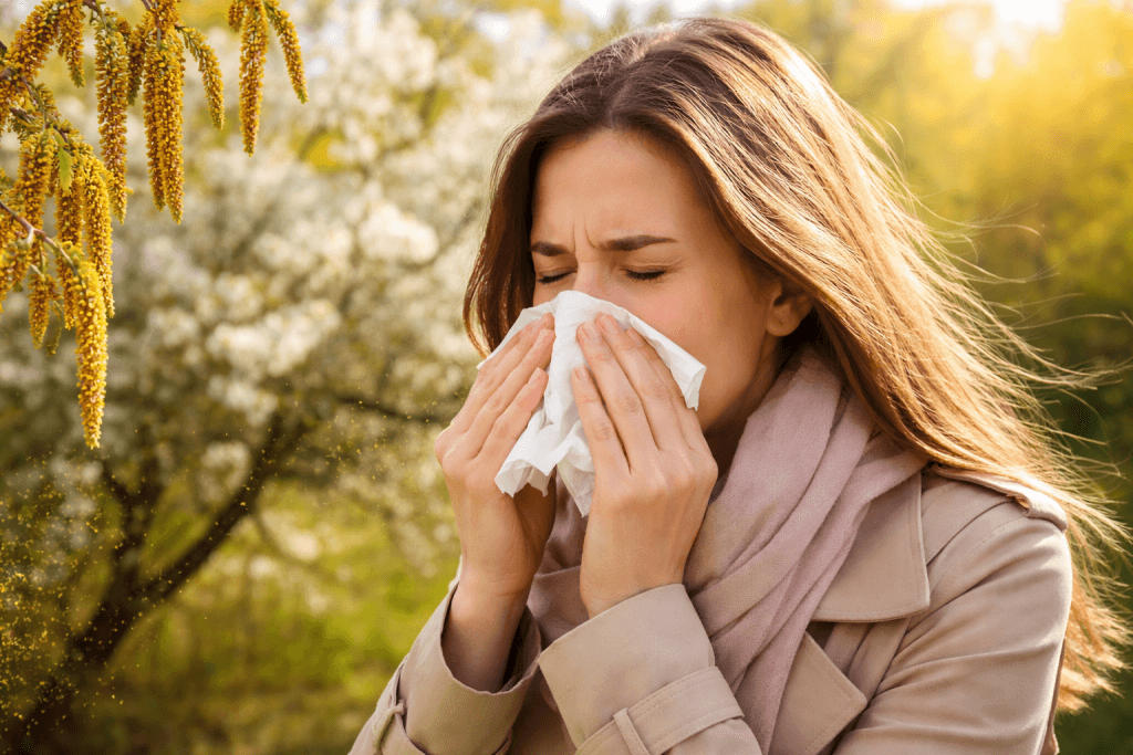 Woman sneezing into a tissue outdoors, with spring blossoms in the background and sunlight.