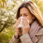 Woman sneezing into a tissue outdoors, with spring blossoms in the background and sunlight.