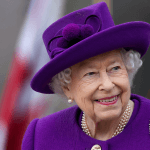 Smiling elderly woman in a purple hat and coat, wearing pearls and a brooch outdoorsan elegant royal-like look (no identity implied)