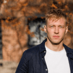 Portrait of a young man with blond hair wearing a white T-shirt and a navy shirt, standing outdoors with autumn leaves and a brick building behind him.