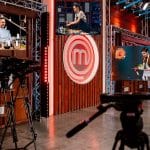 Behind-the-scenes MasterChef studio with multiple cooks on screens and professional cameras filming the competition area.