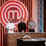 Three MasterChef judges stand behind a wooden judging table on a set, with a large red MasterChef emblem in the background.