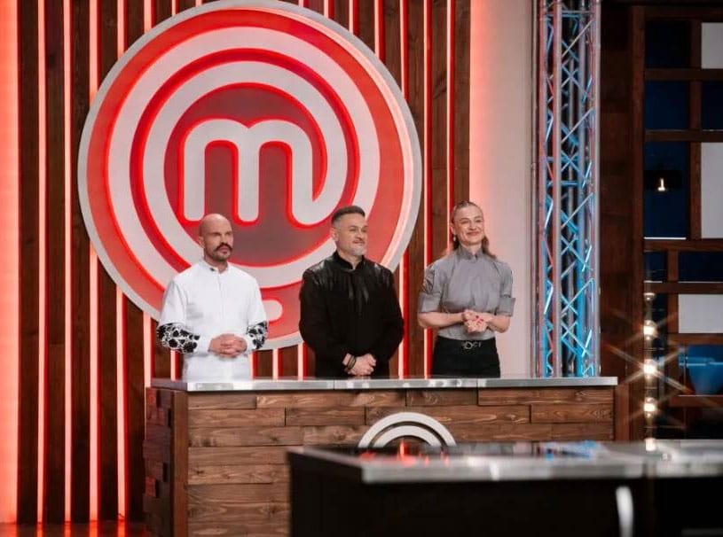 Three MasterChef judges stand behind a wooden judging table on a set, with a large red MasterChef emblem in the background.