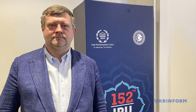 Smiling man in a blue blazer and white shirt stands beside a blue banner with logos, including the Inter-Parliamentary Union, and the text '152nd IRU'.