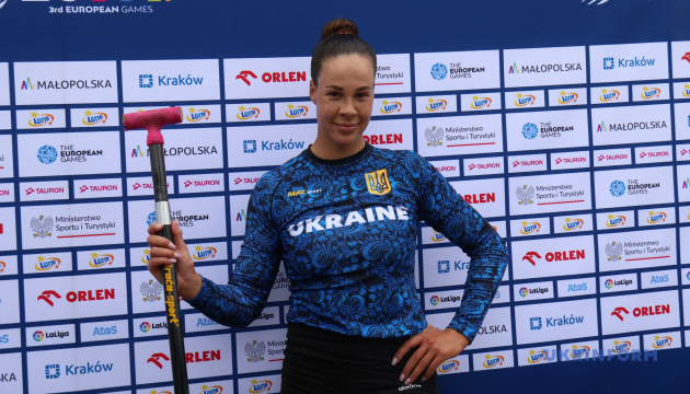 Ukrainian female athlete in a blue Ukraine jersey stands in front of a sponsor backdrop, holding a hammer with her right hand and resting her left hand on her hip.
