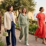 Group of women in stylish outfits walking along a seaside garden path with flowers and a stone wall by the ocean.