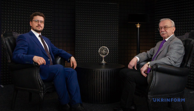 Two men in suits sit in leather chairs in a recording studio with a small round table and a globe centerpiece between them, suggesting a podcast or interview setting.