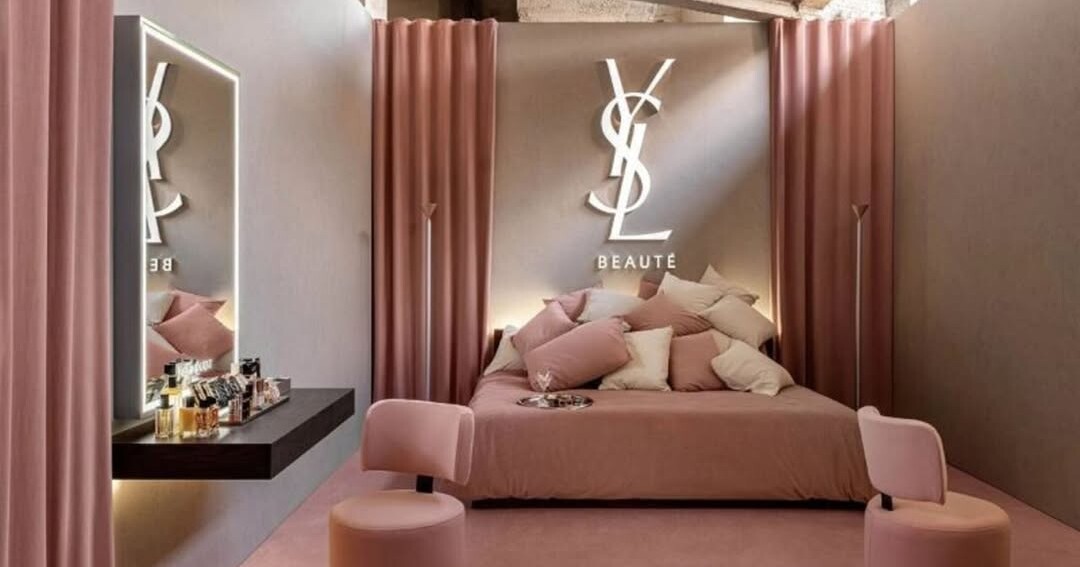 A pink themed boutique room with a bed, plush pillows, and the YSL Beauté logo on a wall behind the bed.