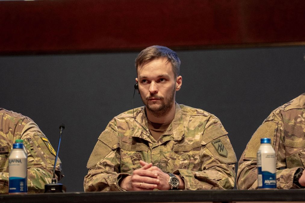 Soldier in camouflage with headset at a panel table, hands clasped in front of a microphone.