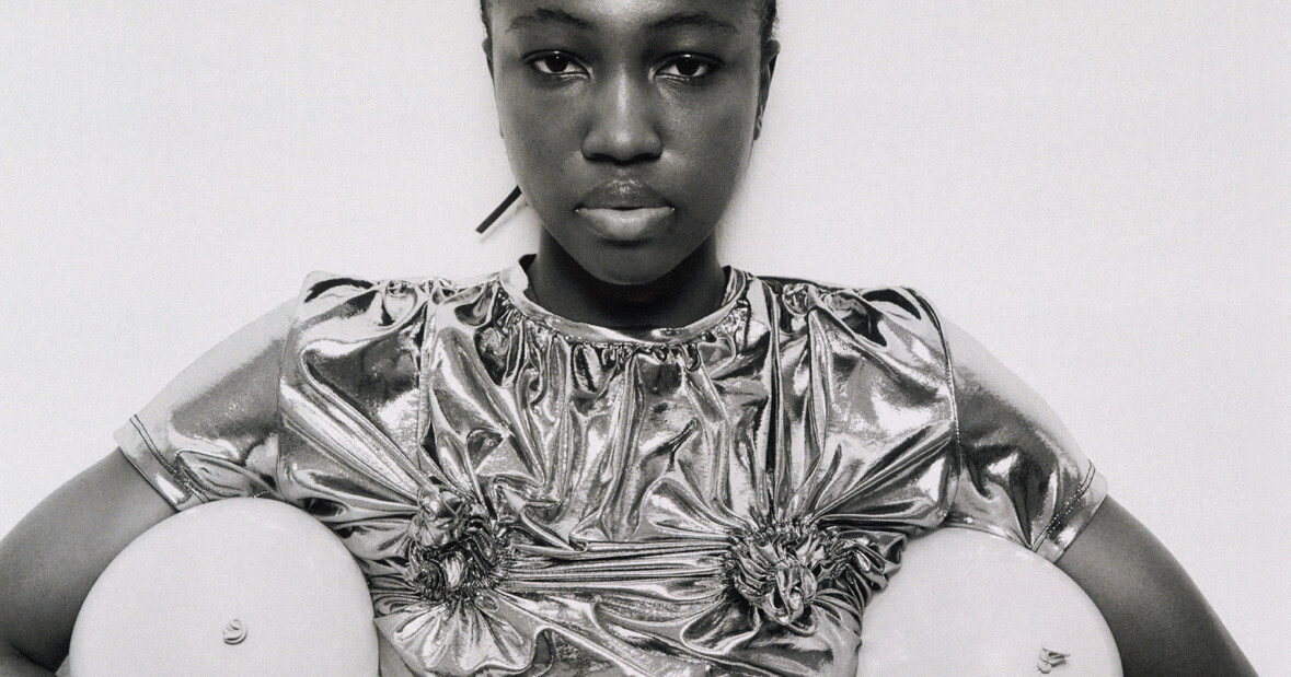 Portrait of a woman in a shiny metallic top, facing the camera with round objects at her sides.