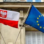 ноутбук, ручка, горнятко кави та маленька польська прапорець-пін на простому тлі